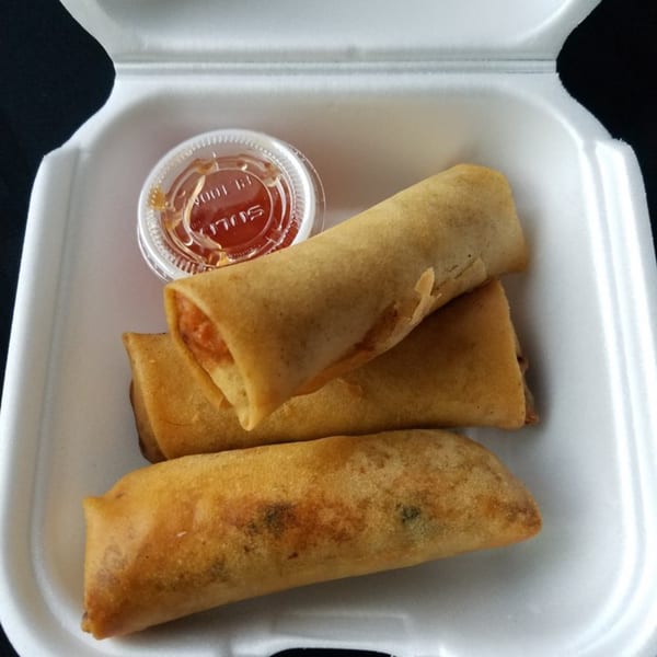 Three golden-brown Yummy Deli spring rolls and a small container of dipping sauce in a white takeout box.