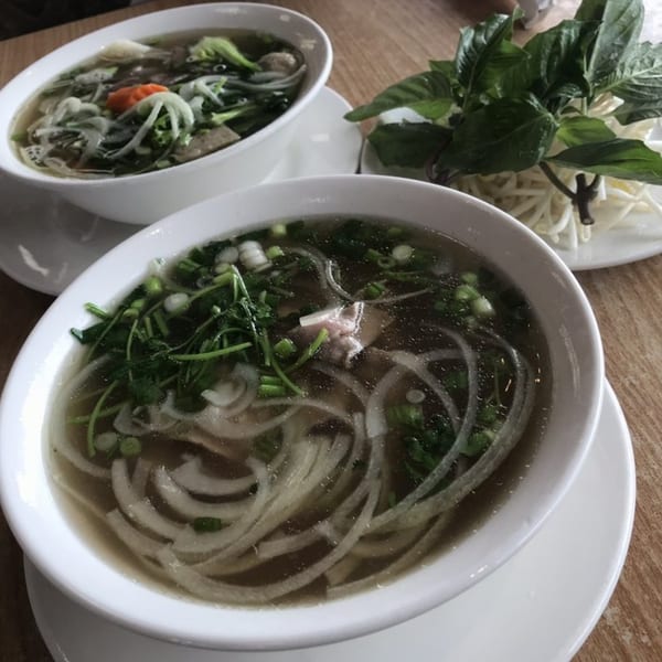 Two bowls of pho with noodles, meat, and vegetables, alongside a plate of fresh herbs.