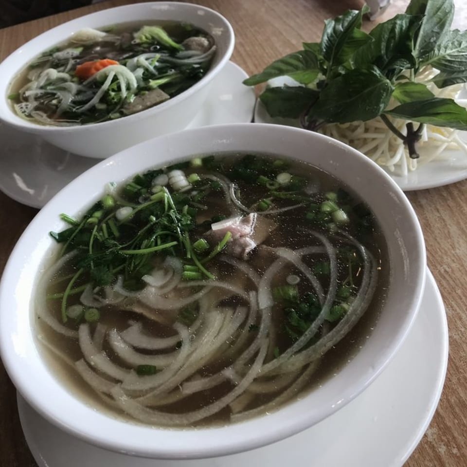 Two bowls of pho with noodles, meat, and vegetables, alongside a plate of fresh herbs.