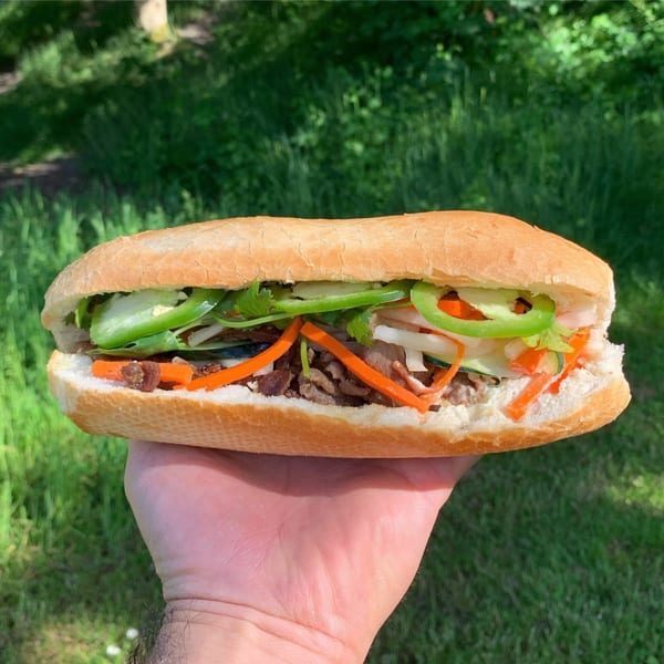 A hand holds a Yummy Deli sandwich filled with meat, carrots, and green peppers against a grassy background.