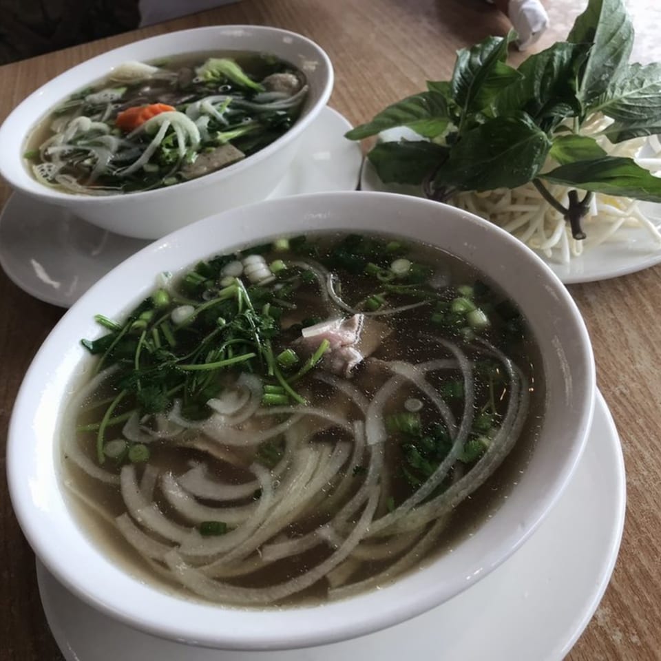 Two bowls of pho with noodles, meat, and herbs, alongside a plate of fresh basil and bean sprouts.